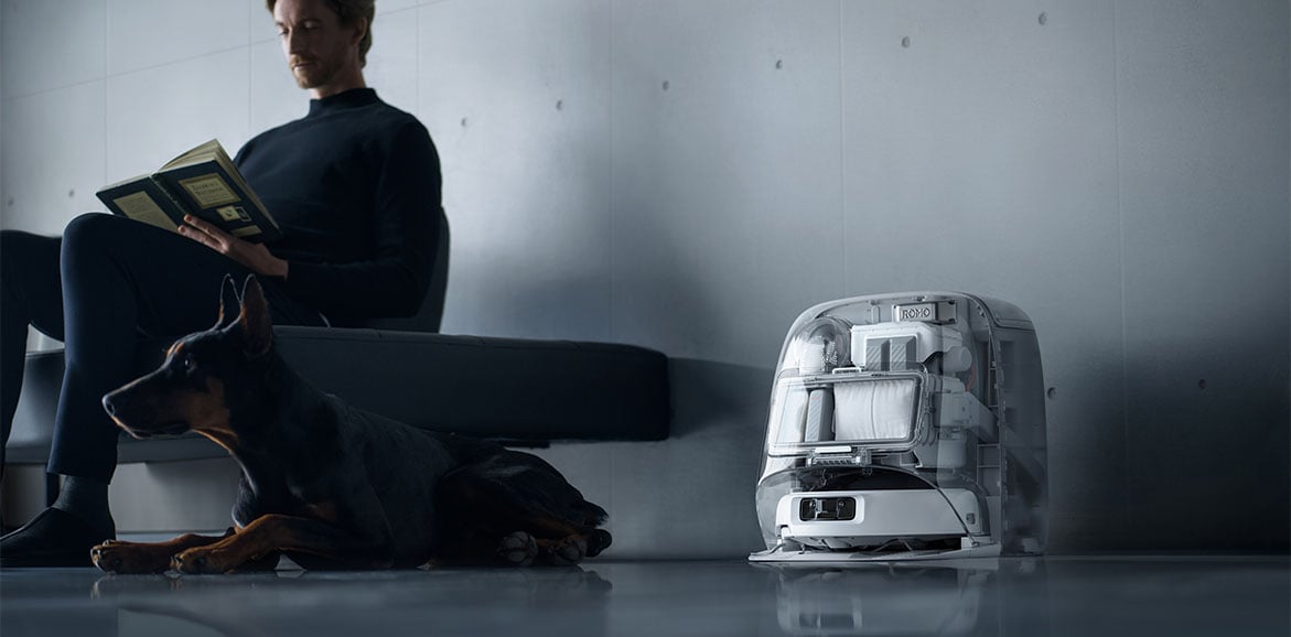 Robot sprzątający DJI ROMO P w stacji dokującej stoi obok kanapy na której siedzi mężczyzna